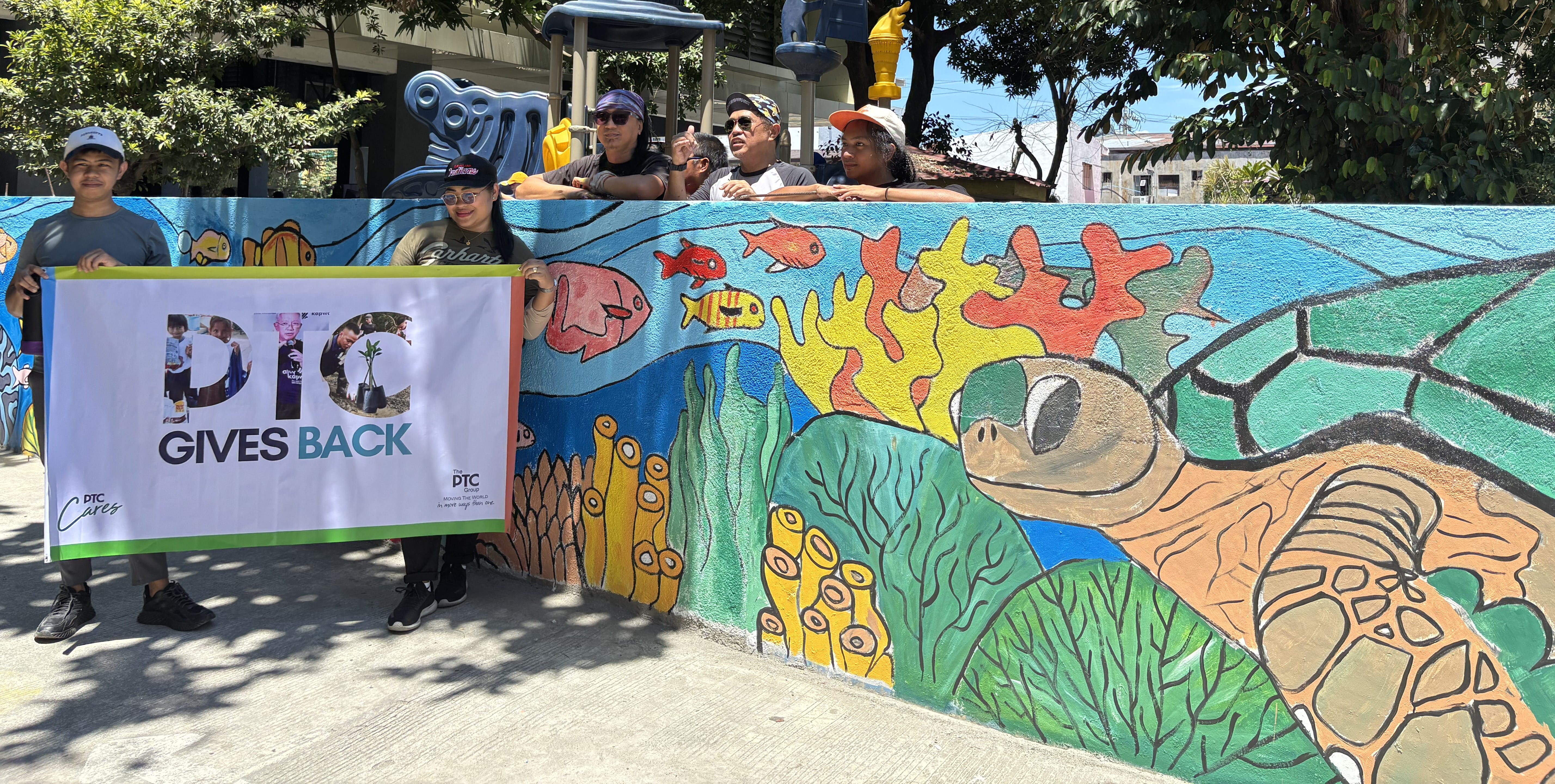 Two volunteers holding a 'PTC Gives Back' banner in front of a colorful marine mural featuring a sea turtle.
