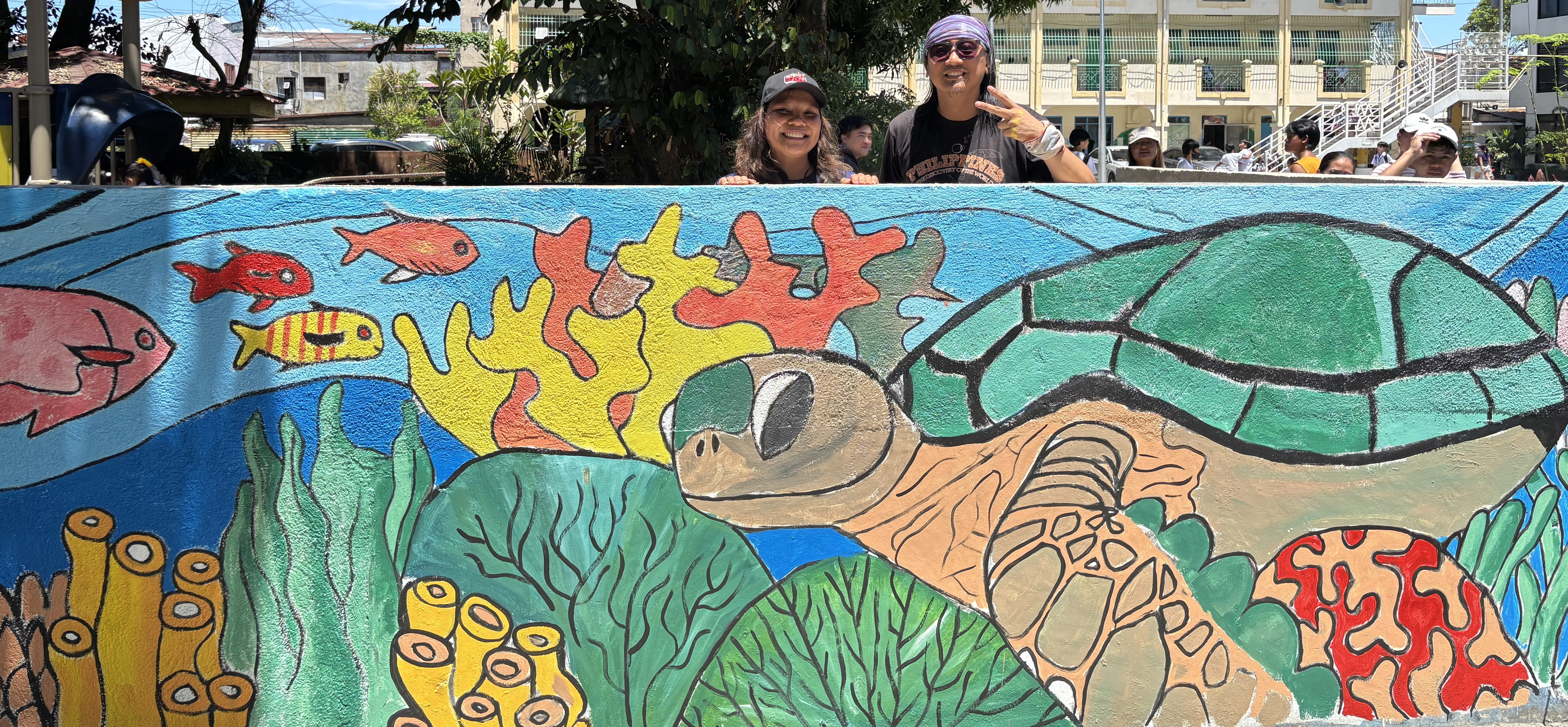 Two volunteers smiling over the top of the wall featuring a large green sea turtle and vibrant orange corals.